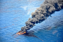 An aerial view of fire burning on a body of water with a large black smoke stack rising from it. 