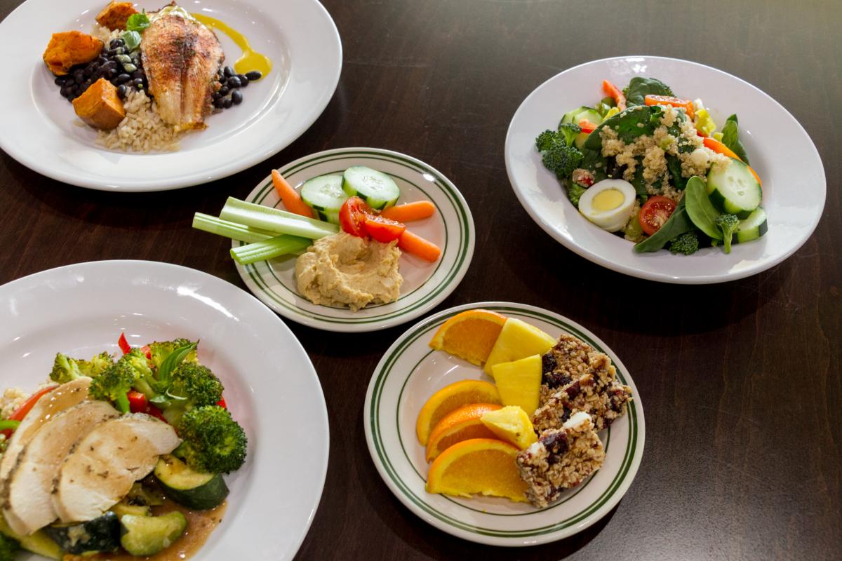 An array of dishes including: quinoa salad; sliced chicken breast with gravy and seasonal vegetables; tilapia over sweet potatoes, black beans and rice; and fresh orange slices with sweet granola energy bars.