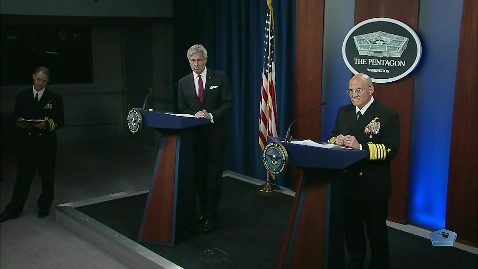 A civilian and a Navy admiral stand at lecterns.