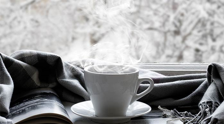book, scarf, and steamy mug on a table facing a window with a snowy landscape outside