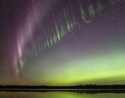 A photo showing a purple arc of light alongside a green picket fence like structure in the night sky above a body of water