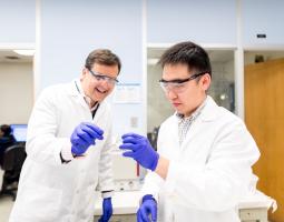 2)	Two men wearing lab coats, protective eyewear, and surgical gloves each hold one end of a long, slender tube.