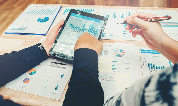 hands holding tablet and papers on desk show graphs and charts