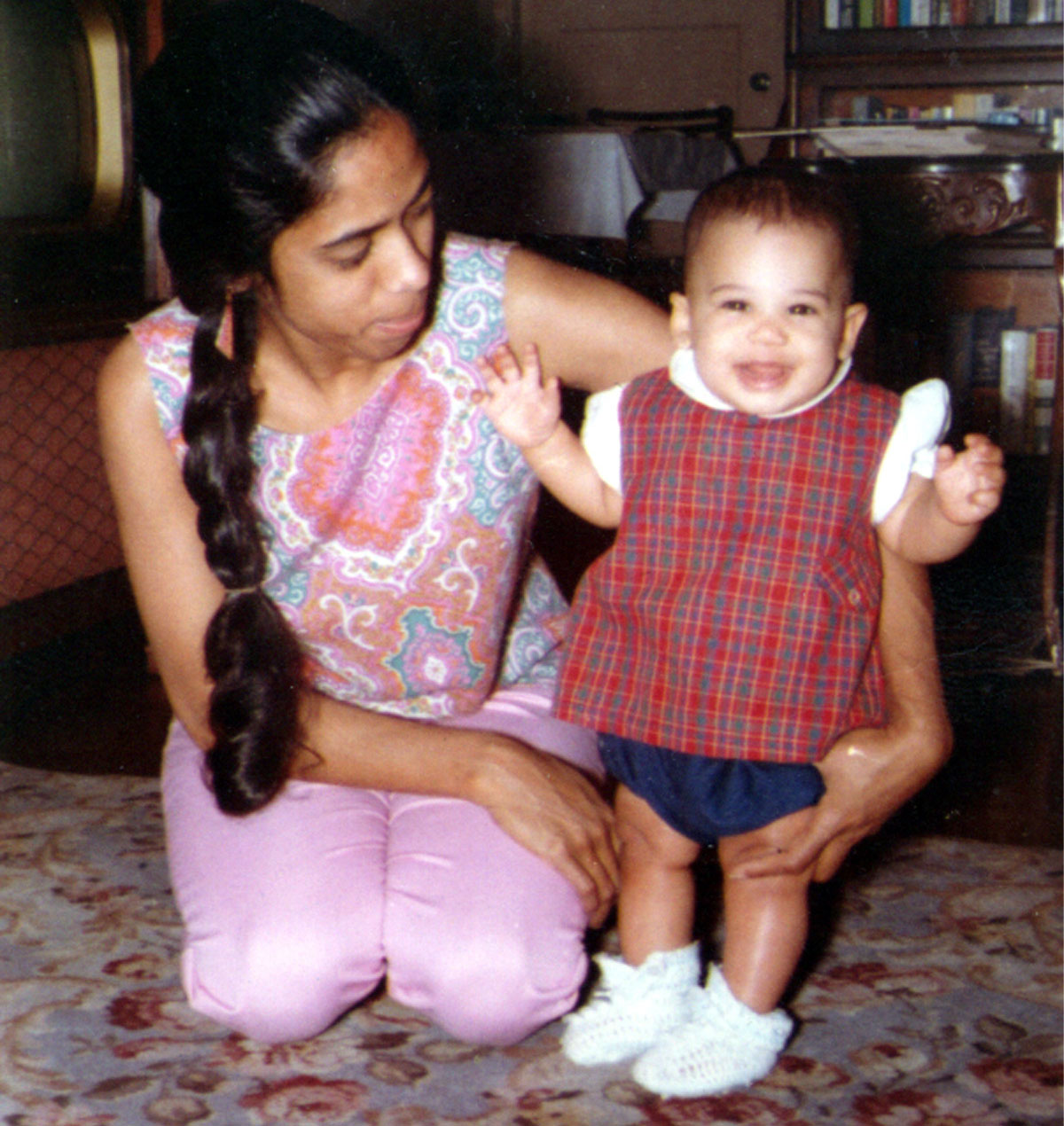 A toddler-aged Kamala smiles with her mother, Shyamala, in front of a bookshelf.