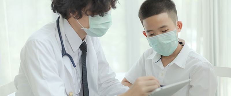 Doctor and boy patient talking with masks while doctor is writing on a tablet.