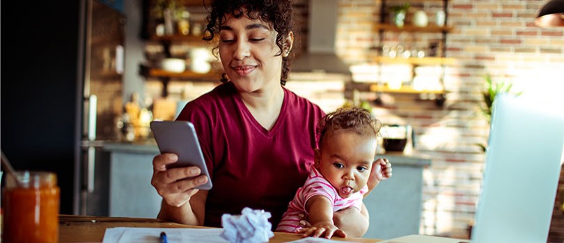 Woman smiling and using her phone while holding her baby.