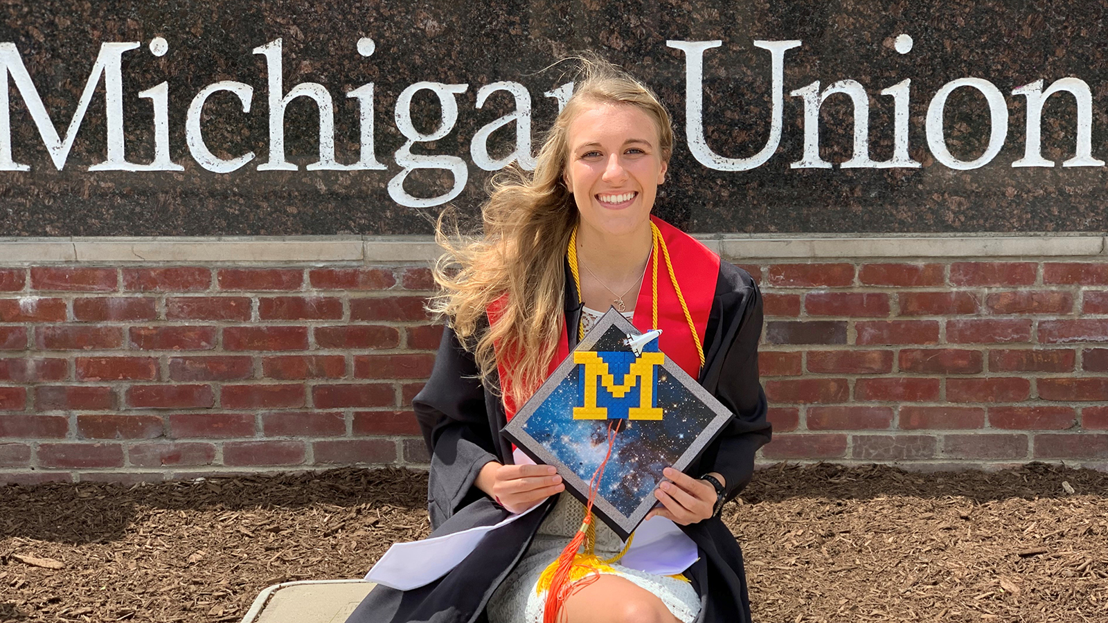 Kaelan Oldani wears her graduation gown and holds her cap while posing in front of a sign that reads 'Michigan Union.'