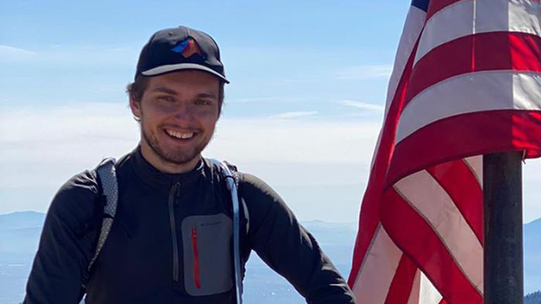 Lucas Lange wears hiking gear and poses next to an American Flag at the top of a mountain with a valley visible in the background.