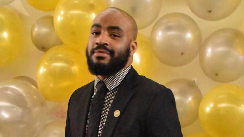 Yohn Ellis, wearing a suit and tie, poses in front of yellow and gold balloons.