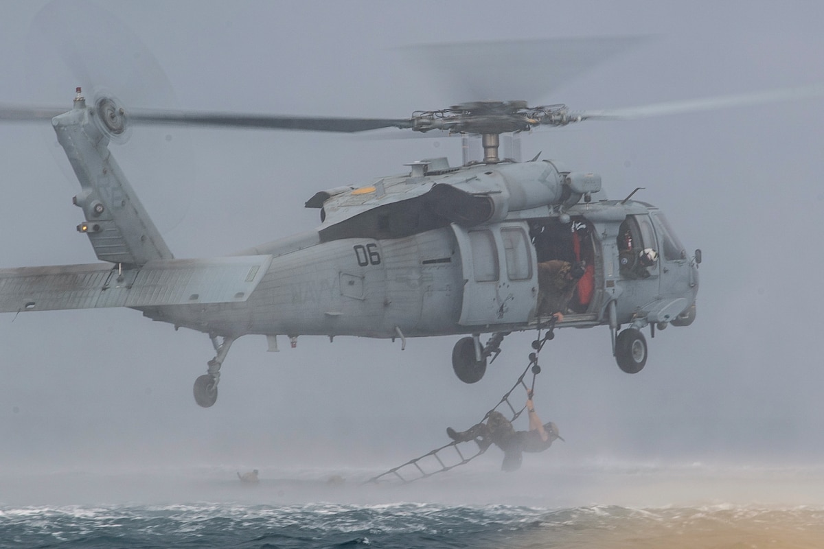 A sailor climbs on a ladder hanging out of an open helicopter hovering over water.