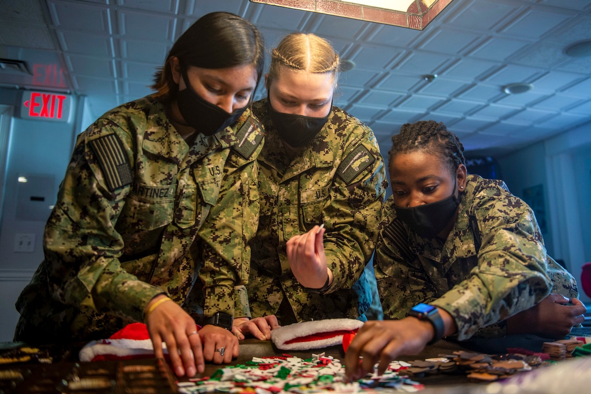 Three sailors in face masks reach for decorations spread out on a table by Christmas stockings.