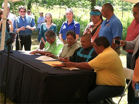 Four men, sitting at a table in front of a crowd of onlookers, sign the deed for Hall Mountain.