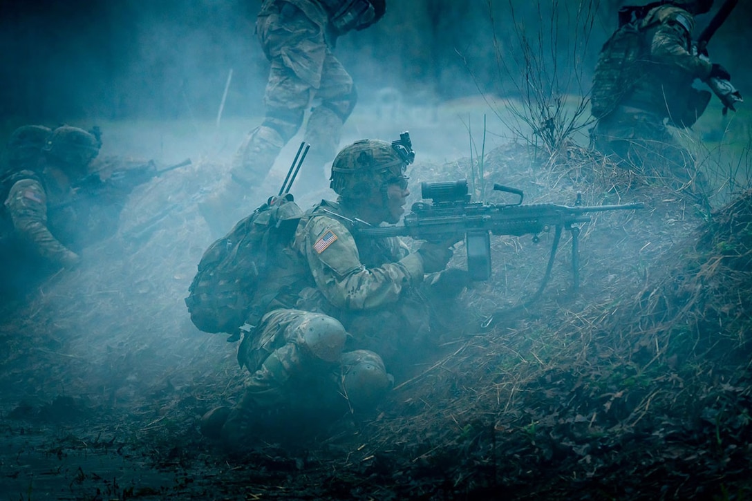 A soldier sits behind a hill and aims a weapon amid blueish smoke.