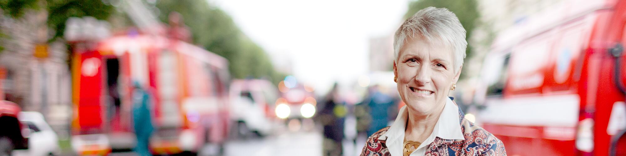 A woman smiling in front of a blurred image of firemen