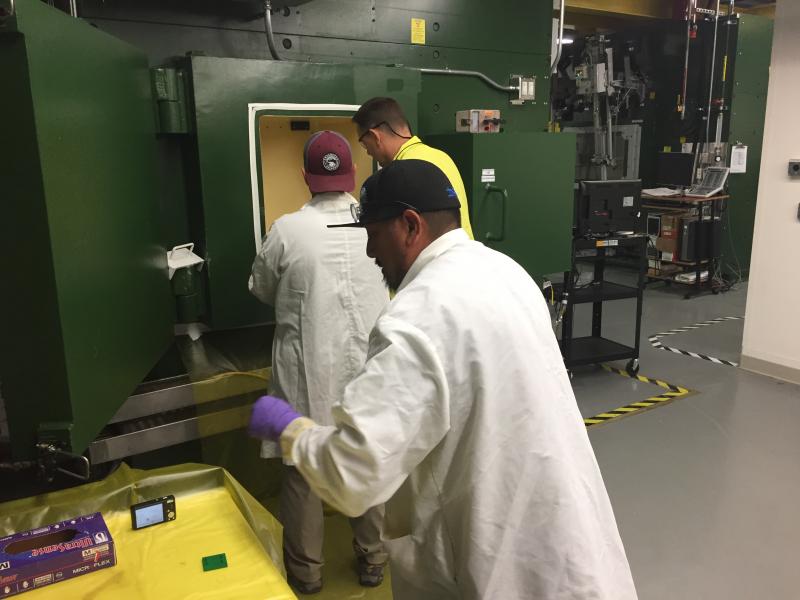 Three men standing outside hot cell, looks like a large green metal box with a window