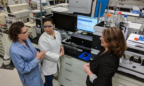 Carolyn Burdette, Laura Regalado and Katrice Lippa having an animated discussion in the lab. Behind them is a computer screen and other laboratory equipment, including a liquid chromatography mass spectrometer. 