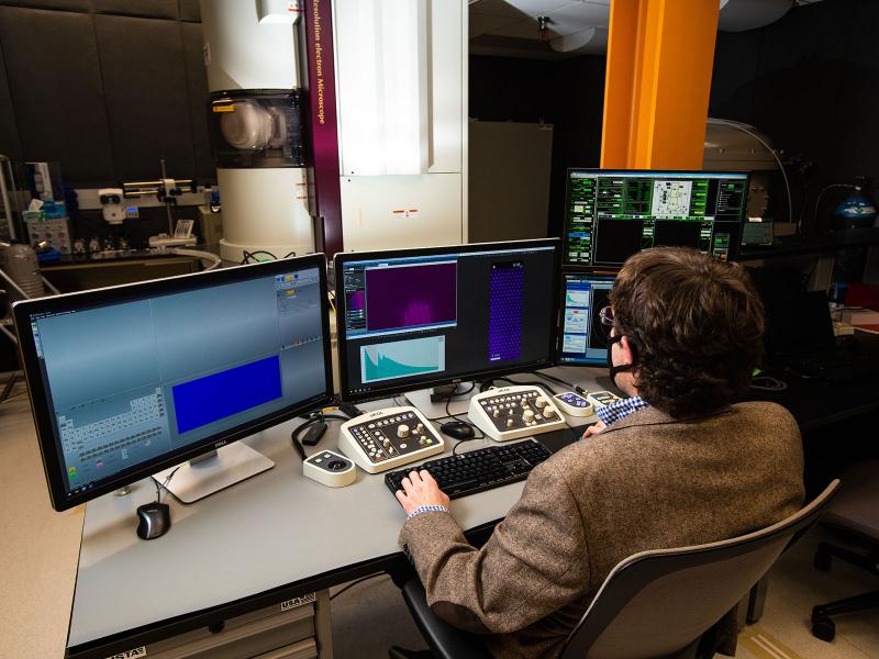 Scientist Steven Spurgeon sits at a desk with a keyboard and multiple monitors while he controls a huge microscope in the room.