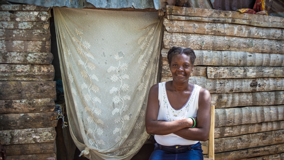 A smiling Haitian woman stands next to the doorway of her home.