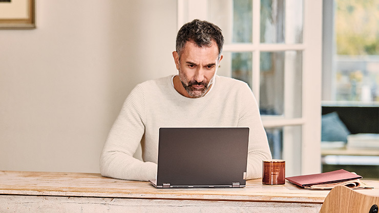 Man uses a laptop computer at home while seated at a long wooden table.