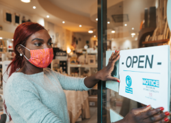 A woman wearing a mask hangs an "open" sign on the door of her business.
