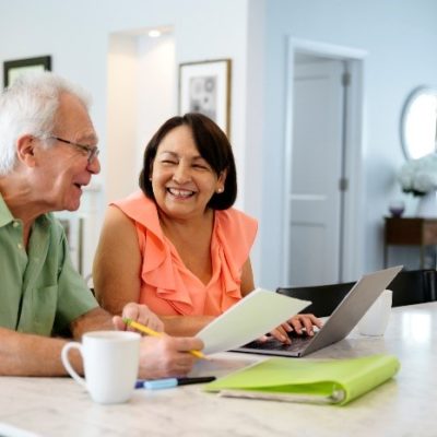 An eldery couple smiling at each other while reviewing finances on a laptop.