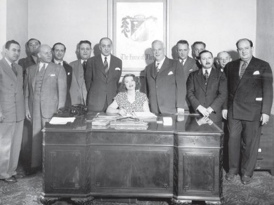 woman at a desk with group of men standing behind her.