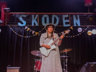 Woman performing on stage with guitar, SKODEN banner behind her