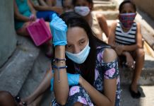 Mulher usando máscara e luvas sentada entre um grupo de pessoas em uma área externa (© Ariana Cubillos/AP Images)