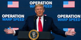 Donald Trump standing at lectern with outstretched arms in front of Operation Warp Speed background (© Evan Vucci/AP Images)