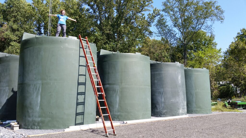 Woman standing with outstretched arms on one of four giant grey tanks, with an orange ladder propped up beneath her.