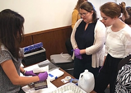 Three women stand around a table with a rectangular aluminum tray on it. One woman holds a booklet in her purple-gloved hands while the two other women look at her.