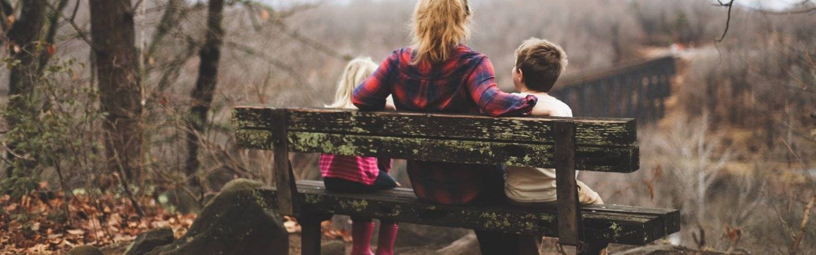 Photo credit: Benjamin Manley. A woman wearing flannel sits on a bench with two young children, back to the viewer.