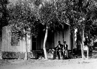 Two workers rest on the porch of a small house surrounded by trees.