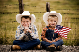 Two boys in cowboy hats sitting on country road with U.S. flag. (iStock)