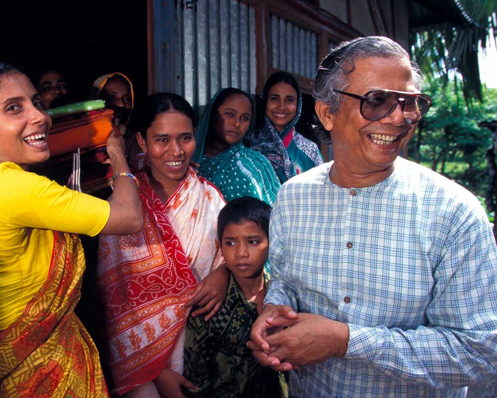 Muhammad Yunus in sunglasses looks to the right and smiles, surrounded by smiling people in a doorway.