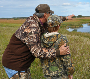 An older gentleman points over a wetland with a young hunter at his side.
