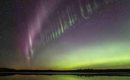 A photo showing a purple arc of light alongside a green picket fence like structure in the night sky above a body of water