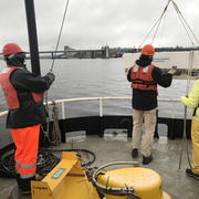 3 people on a boat lowering an instrument into the water on a pulley