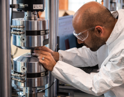 Man attired in lab clothing and classes working with his hands on a metal cylinder.