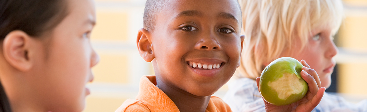 Boy smiling and eating an apple in school lunch room.