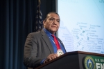 Director Frost stands at a podium with the DOE seal; behind him is a US flag.