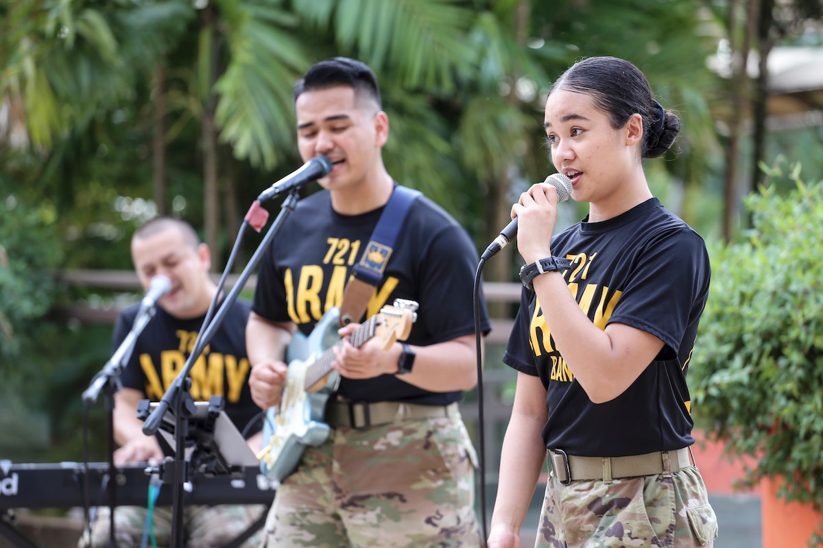 Three soldiers sing into microphones in a tropical outdoor setting; one also plays guitar and one plays keyboard.