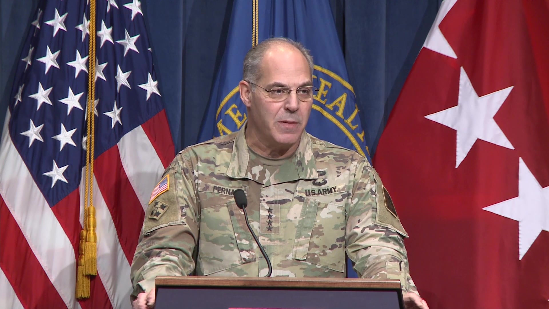 A soldier speaks at a lectern in front of flags.