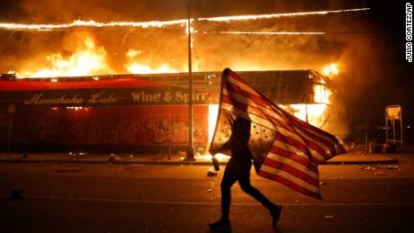 A protester carries a U.S. flag upside down, a sign of distress, next to a burning building Thursday, May 28, 2020, in Minneapolis. Protests over the death of George Floyd, a black man who died in police custody Monday, broke out in Minneapolis for a third straight night. (AP Photo/Julio Cortez)