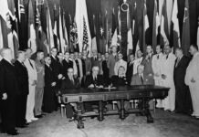 Men seated at table with others surrounding them, with various flags behind them (© Bettmann/Getty Images)