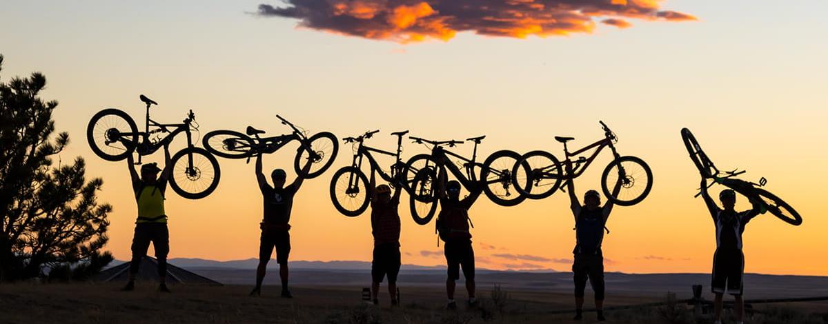 Six e-bike riders are silhouetted against a sunset as they all hold their bikes above their heads