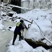 person in coat and hat digging with shovel on snow and ice covered stream by frozen waterfall 