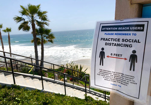 Imagen de una playa a la distancia. Un afiche en primer plano que informa que las personas deberían mantener al menos 6 pies de distancia entre sí mientras usan las playas.