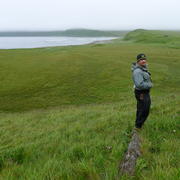 A man standing on a log in the grass near a bay