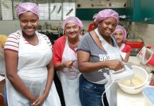 Women in aprons smiling in kitchen, with one using hand mixer in bowl (© Geovanni Hernandez/AES)